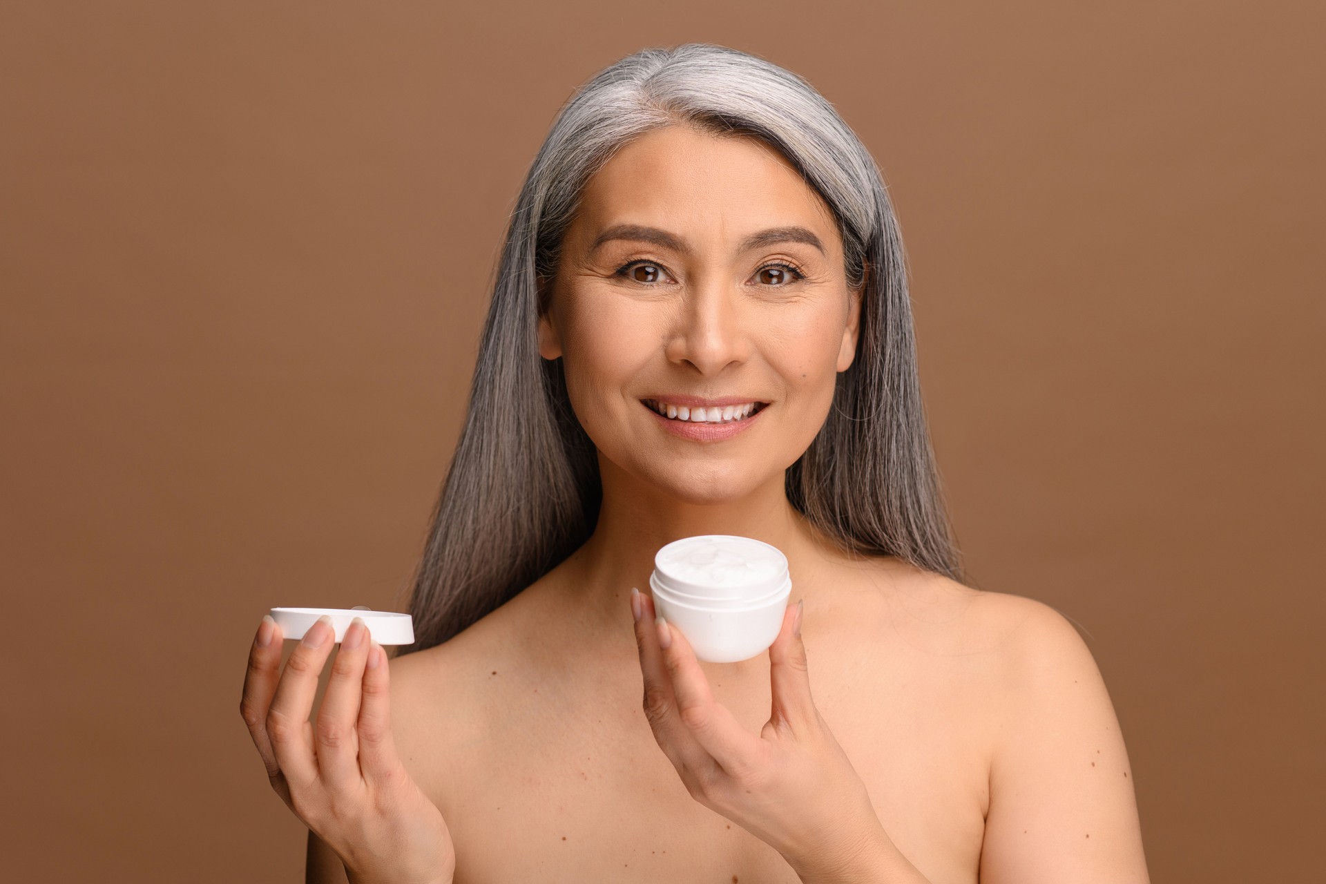 Serene middle-aged Asian woman with fresh clean healthy skin stands with jar of moisturiser anti-aging cream